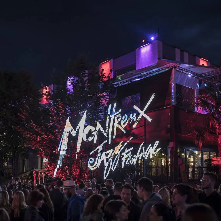 Menschenmenge vor beleuchtetem Gebäude mit "Montreux Jazz Festival"-Projektionsschriftzug.