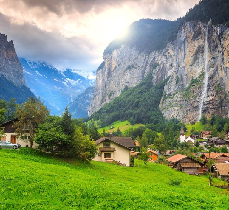 Berglandschaft mit Wasserfall, umgeben von grünen Wiesen und malerischen Häusern.