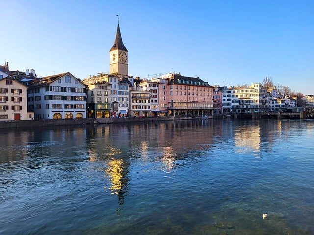 Häuserzeile am Fluss mit einem Kirchturm im Hintergrund bei klarem Himmel.