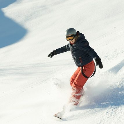 Person in schwarzer Jacke und orangefarbener Hose fährt Snowboard im Schnee.